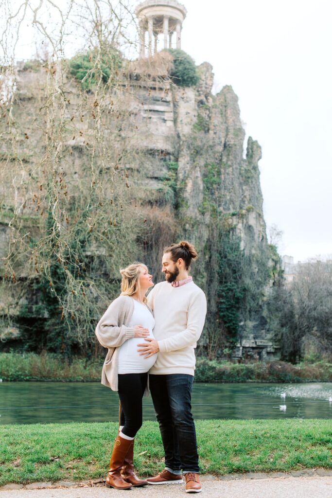 pregnant couple standing in front of buttes chaumont in paris